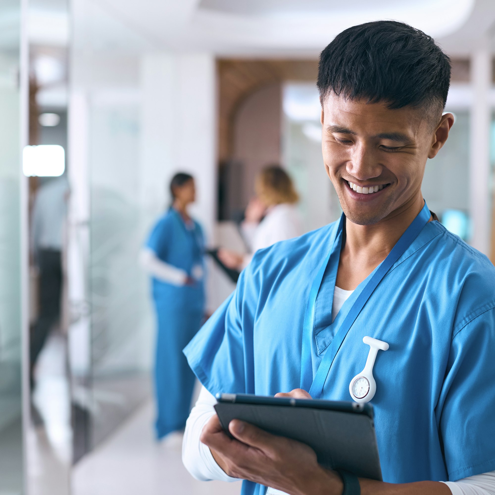 Male Doctor Or Nurse Wearing Scrubs With Digital Tablet Checking Patient Notes In Hospital